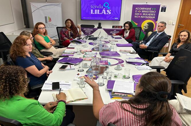 Foto colorida: reunião em mesa com materiais da campanha Tenda Lilás, sala institucional, enfrentamento à violência, debatendo ações em roxo, branco e verde