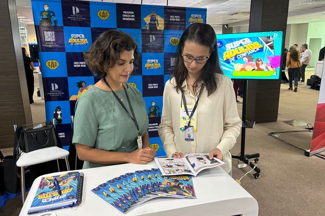 Foto colorida: duas mulheres folheiam cartilhas informativas em estande institucional, ambiente interno, ação educativa, orientando público em azul, branco e verde