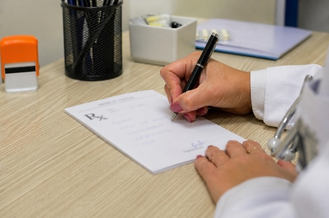Foto colorida: mão escreve receita médica sobre mesa, consultório clínico interno, atendimento de saúde, prescrevendo tratamento em branco, bege e preto