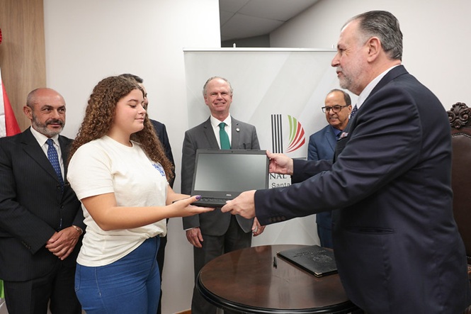 Foto horizontal: ao centro, presidente Rubens Schulz observa desembargador Marcio Rocha Cardoso entregar notebook a jovem, em ato institucional.