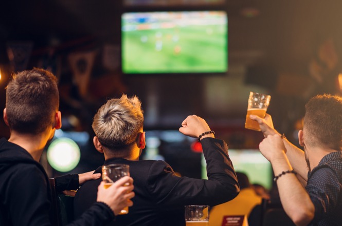 Foto colorida: torcedores erguem copos diante da TV, bar interno, jogo de futebol, comemorando em verde e âmbar