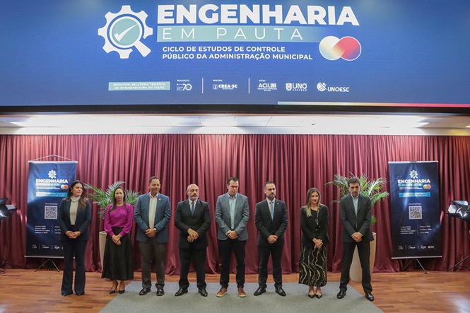 Foto horizontal: desembargador André Luiz Dacol entre autoridades em evento “Engenharia em Pauta”, palco com cortinas, tons vermelho e azul.