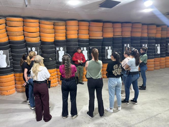 Em um estande de tiro com paredes revestidas por pneus empilhados nas cores preta e laranja, um instrutor vestido com camiseta vermelha orienta um grupo de mulheres posicionadas em semicírculo à sua frente. As participantes usam protetores auriculares e vestem roupas casuais, enquanto observam atentamente as instruções. Ao fundo, diversos alvos de tiro humanoides estão fixados ao longo da parede, caracterizando um ambiente de treinamento prático e supervisionado.
