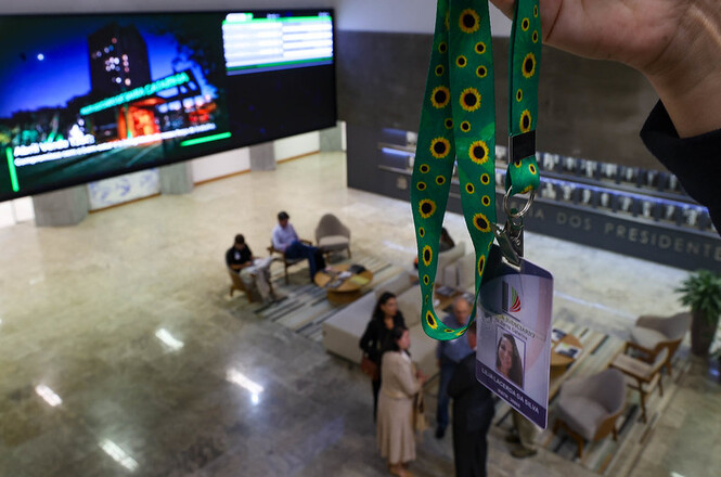 Foto colorida: mão exibe cordão com girassóis diante do prédio do TJSC, área externa institucional, simbolizando inclusão em verde e amarelo