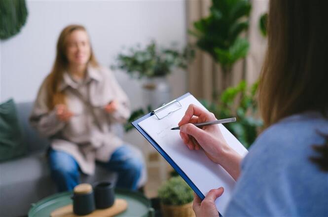 Foto colorida: profissional anota em prancheta enquanto pessoa fala no sofá, consultório interno, escuta institucional, registrando relato em verde, bege e branco