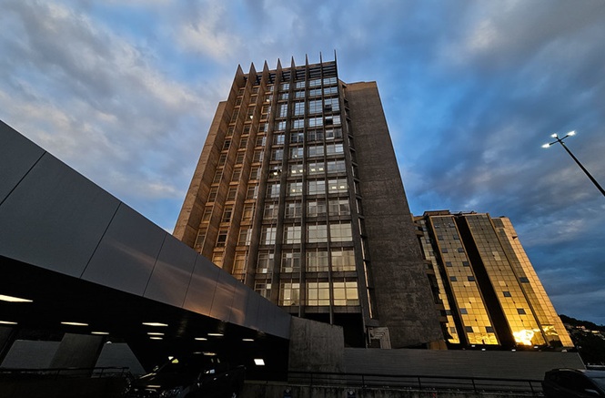 Foto colorida: prédio do Tribunal de Justiça de Santa Catarina ao entardecer, área urbana, sede institucional, destacando arquitetura em cinza, dourado e azul