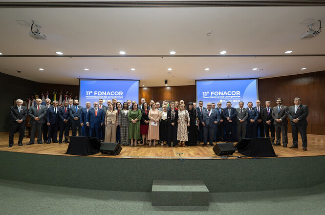 Foto colorida: grupo de autoridades posando em palco, auditório institucional interno, 11º FONACOR, registrando encontro em azul, preto e cinza