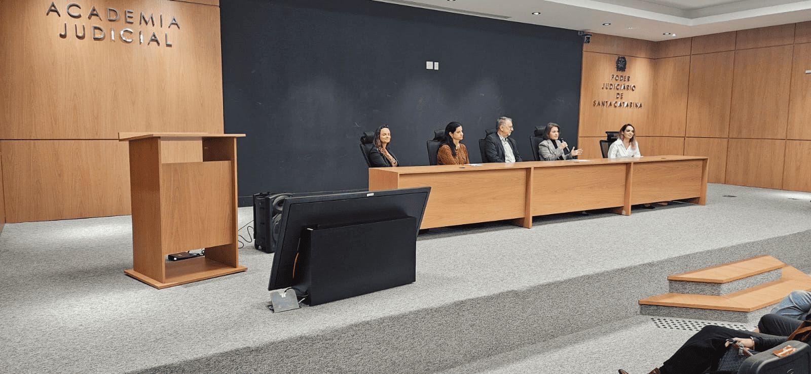 imagem mostra um auditório com palco elevado e paredes revestidas em madeira clara. Ao centro, cinco pessoas estão sentadas atrás de uma mesa longa de madeira durante uma solenidade. À esquerda, há um púlpito vazio e, à frente do palco, um monitor de televisão inclinado no chão. Nas paredes ao fundo aparecem as inscrições “Academia Judicial” e o brasão do Poder Judiciário de Santa Catarina. O ambiente é amplo, com iluminação embutida no teto e parte da plateia aparece no canto inferior direito.