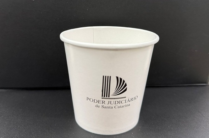 Foto colorida: copo biodegradável com logomarca do Poder Judiciário, fundo neutro, ação sustentável, exibindo design em branco e preto