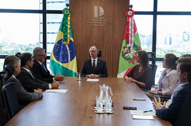 Foto horizontal: presidente do TJSC Rubens Schulz ao centro, entre as bandeiras do Brasil e de Santa Catarina, conduz reunião com autoridades, tons madeira e claros.