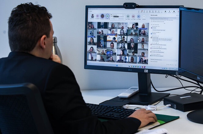 Foto colorida: pessoa diante de monitor com videoconferência, escritório interno, trabalho remoto, participando de reunião em azul, branco e cinza