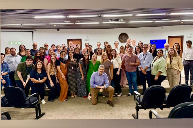 Foto colorida: grupo reunido em auditório institucional, ambiente interno amplo, cerimônia de homenagem, posando para registro em verde, bege e preto