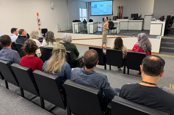 Foto colorida: palestrante diante do público sentado, plenário institucional interno, evento formativo, apresentando conteúdo em branco, cinza e bege