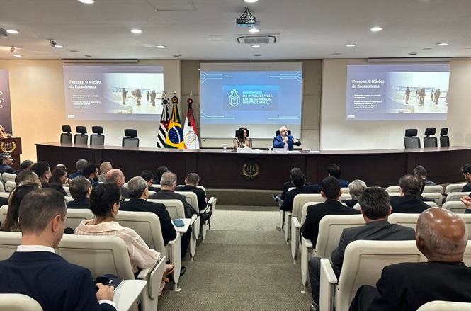 Foto colorida: público e mesa de debatedores em auditório institucional, evento de segurança, acompanhando palestra em azul, branco e cinza