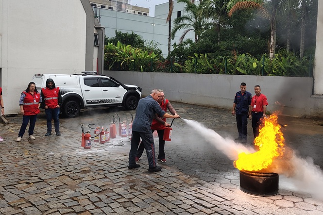 Foto colorida: pessoas usando extintor em pátio externo, treinamento de segurança, combatendo chamas em laranja, vermelho e cinza