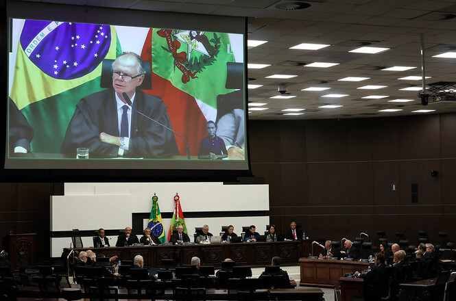 Foto horizontal: no telão, desembargador Rubens Schulz participa de sessão no plenário, autoridades à mesa, tons escuros.