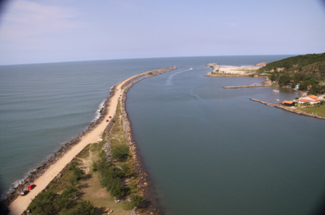 Foto colorida: barra costeira vista do alto, encontro do mar e lagoa, paisagem natural, destacando faixa de areia em azul, verde e bege