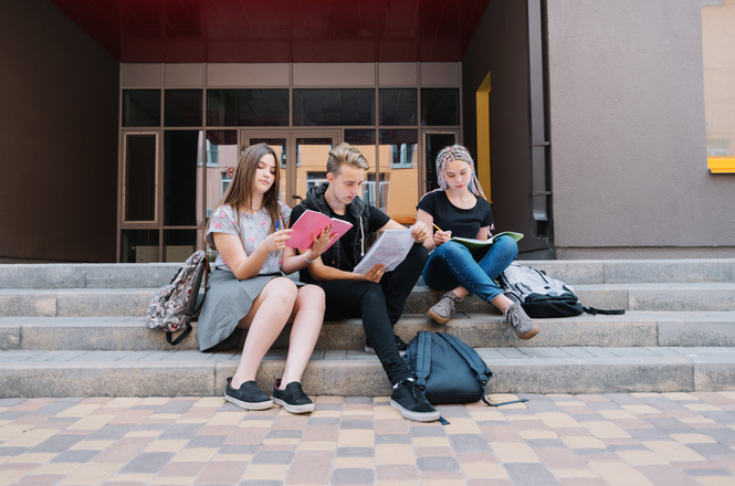 Foto colorida: estudantes sentados em escadaria externa, campus universitário, rotina acadêmica, estudando em azul, bege e cinza