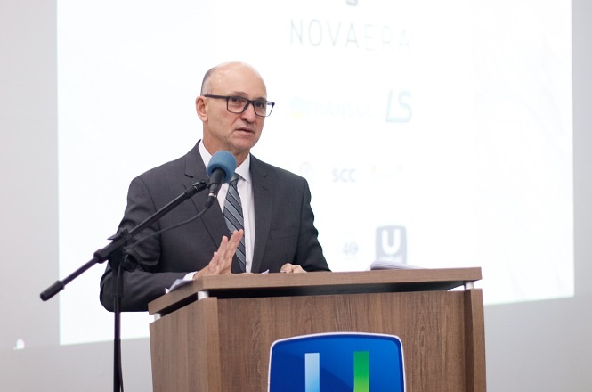 Foto colorida: palestrante ao púlpito, auditório universitário interno, evento acadêmico, apresentando conteúdo em branco, azul e madeira