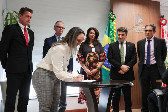 Foto horizontal: mulher assina documento com Rubens Schulz e autoridades em cerimônia no TJSC.