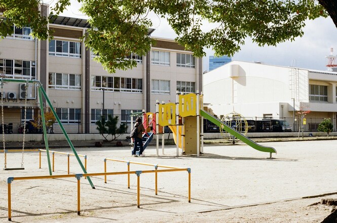 Foto horizontal: parque infantil com escorregador verde e balanços diante de prédio escolar em dia claro.
