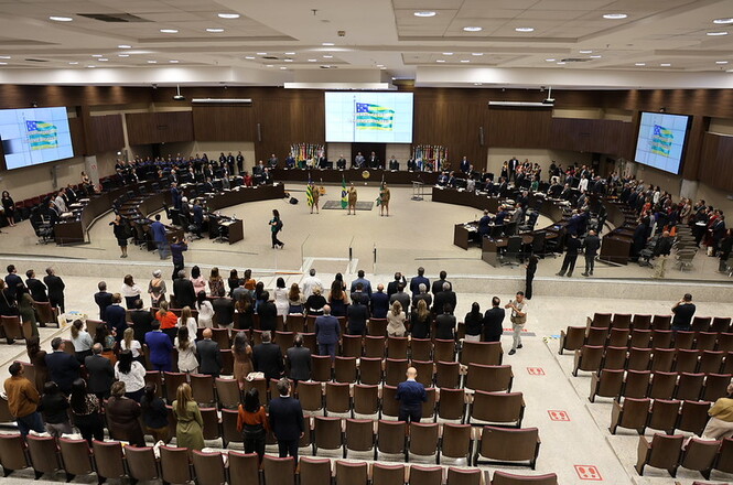 Foto horizontal: plenário com grande público em sessão solene, autoridades dispostas em formato semicircular ao centro, bandeiras ao fundo e participantes em pé, ambiente institucional