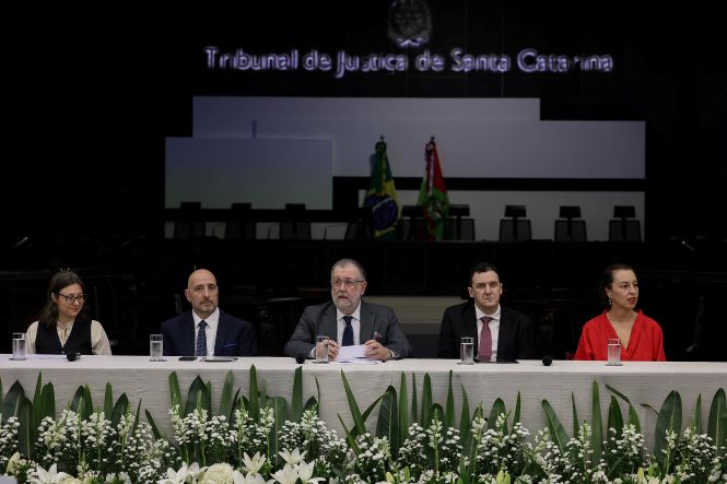 Fotografia mostra cinco integrantes da mesa de abertura do evento sentados atrás de uma longa bancada branca decorada com arranjos de flores brancas e folhagens verdes. Da esquerda para a direita estão Haydee Leppnow, usando óculos e roupa em tons escuros; o juiz Romano Enzweiler, de terno escuro e gravata; o desembargador Márcio Rocha Cardoso, ao centro, de terno cinza e gravata, falando ao microfone; Alexsander Postali, de terno escuro e gravata vermelha; e Carolina Rodrigues Costa, vestindo roupa vermelha. Ao fundo, aparece o palco do auditório do Tribunal de Justiça de Santa Catarina, com cadeiras vazias, as bandeiras do Brasil e de Santa Catarina e a inscrição “Tribunal de Justiça de Santa Catarina” iluminada na parede.