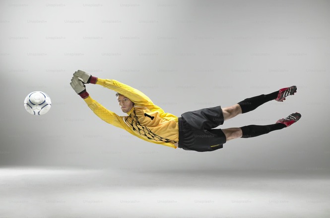 Foto horizontal: goleiro de uniforme amarelo salta para defender bola em fundo branco.