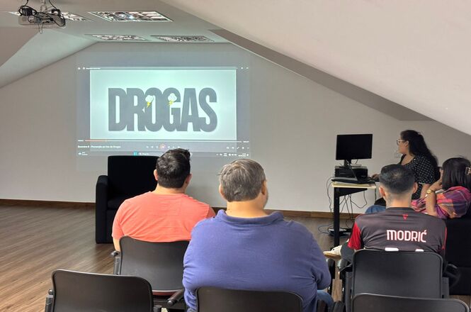 Foto horizontal: pessoas assistem palestra sobre drogas em sala com projeção e cadeiras pretas.