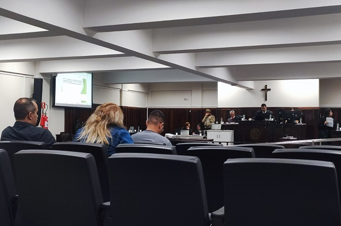 Foto horizontal: audiência ocorre em plenário judicial com público sentado e telão ao fundo.