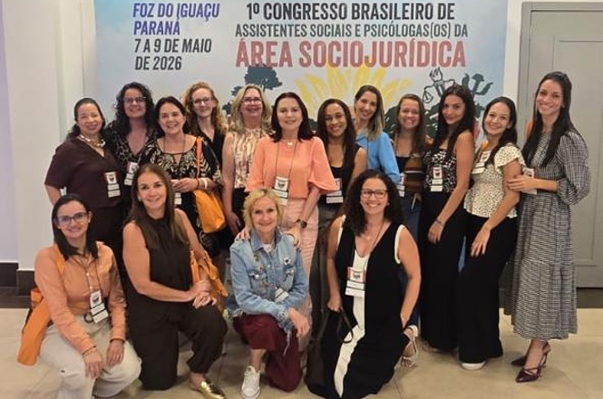 Foto horizontal: grupo de mulheres posa em congresso sociojurídico diante de painel institucional.