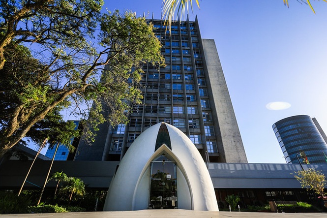 Foto horizontal: capela branca em frente ao prédio do TJSC sob céu azul e árvores verdes.