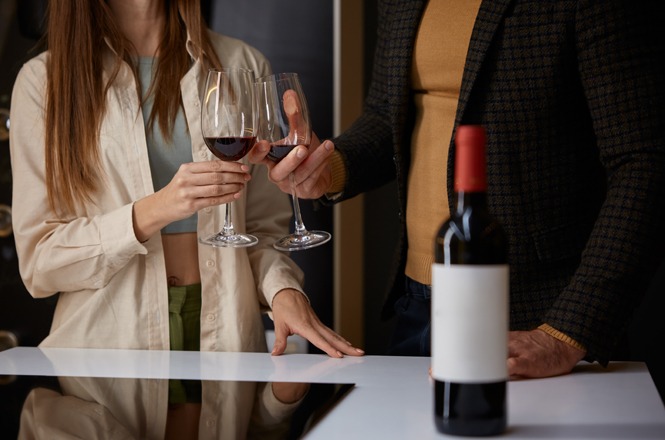 Foto horizontal: duas pessoas brindam com taças de vinho em ambiente interno, garrafa em primeiro plano, tons quentes.