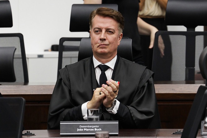 Foto horizontal: desembargador João Marcos Buch aplaude em sessão, traje togado, ambiente institucional, tons neutros.