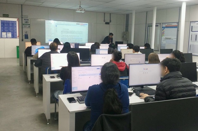 Foto horizontal: alunos utilizam computadores em sala de informática, atividade em aula, professor à frente, tons neutros.