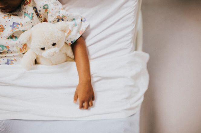 Foto horizontal: criança deitada em leito hospitalar segura urso de pelúcia, mão à mostra, ambiente claro, tons suaves.