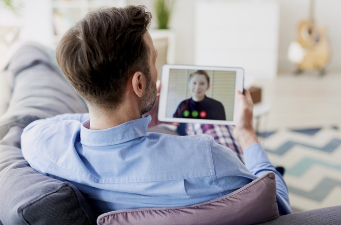 Foto horizontal: homem em casa realiza videochamada em tablet, conversa online em ambiente doméstico, tons claros.