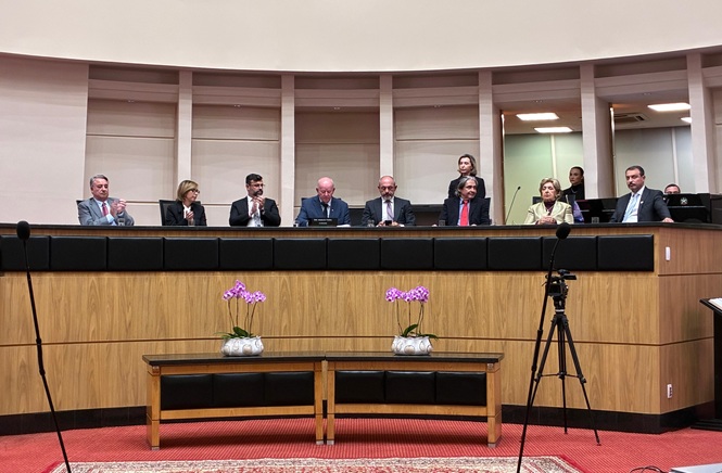 Foto horizontal: autoridades compõem mesa em sessão solene, aplausos em andamento, ambiente institucional, tons madeira e neutros.