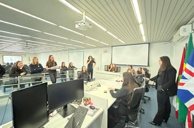Foto horizontal: grupo de mulheres interage em sala de informática, dinâmica em andamento, ambiente institucional, tons claros.