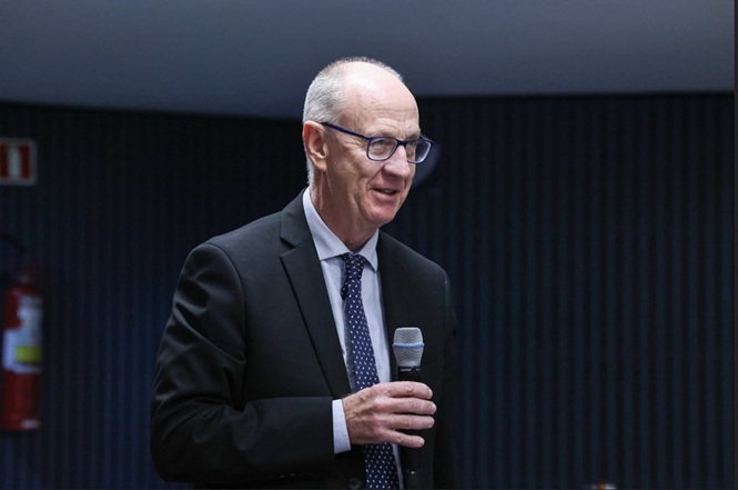 Foto horizontal: homem fala ao microfone em evento, expressão sorridente, ambiente interno, tons escuros.