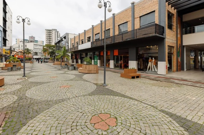 Foto colorida: rua com lojas e calçada em mosaico, área urbana comercial, cotidiano citadino, circulando em cinza, bege e marrom