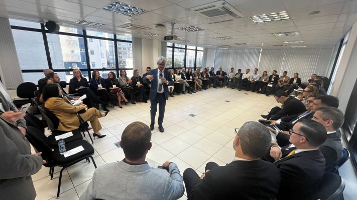 Sala ampla e bem iluminada, com piso claro e paredes brancas, equipada com janelas grandes ao fundo que deixam entrar luz natural. No teto, há luminárias retangulares e aparelhos de ar-condicionado. Cadeiras estão dispostas em formato de círculo, ocupadas por cerca de 30 a 40 pessoas vestidas com roupas formais, como ternos e blazers. No centro do círculo, o desembargador Roberto Bacellar, vestindo terno escuro e camisa branca, segura um microfone e faz uma apresentação ou fala, sendo o foco da atenção do grupo. Outras pessoas ao redor estão sentadas, algumas com blocos de anotação ou dispositivos eletrônicos nas mãos.