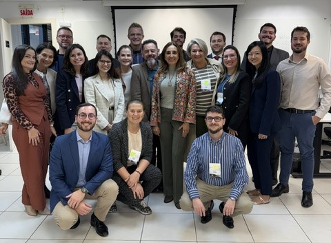 Foto em ambiente interno com cerca de 18 pessoas posando em grupo diante de uma tela de projeção. As pessoas estão organizadas em três filas (algumas agachadas na frente e outras em pé atrás), usando roupas formais ou semiformais e crachás. O espaço é bem iluminado, com piso claro e uma placa de “SAÍDA” visível ao fundo.