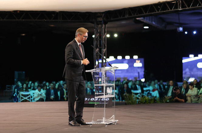 Foto horizontal: desembargador Marcos Probst discursa em púlpito durante evento, público ao fundo, ambiente amplo, tons escuros.