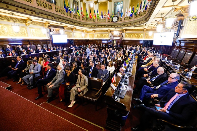 Foto horizontal: plenário histórico lotado durante evento institucional, autoridades e público distribuídos em ambiente solene, tons dourados e madeira.