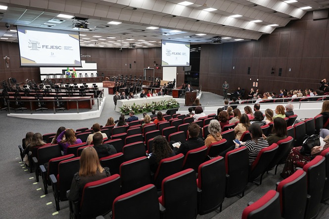 Foto horizontal: auditório recebe evento institucional com público atento, palestrante ao púlpito e projeções ao fundo, tons neutros.