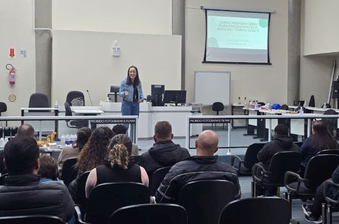 Foto horizontal: mulher apresenta conteúdo em sala de audiência para público sentado, projeção ao fundo, ambiente institucional.