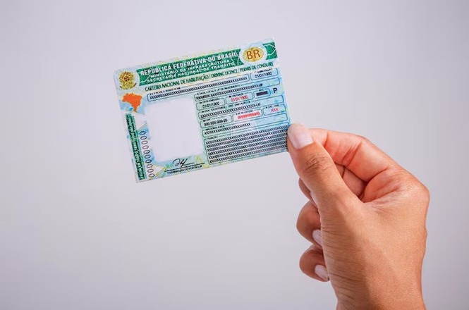Foto mão segurando CNH verde e branca, fundo cinza claro, documental, exibindo documento brasileiro