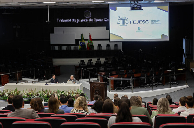 Foto horizontal: auditório do TJSC recebe evento da FEJESC, público em cadeiras vermelhas e painel projetado ao fundo, ambiente institucional.