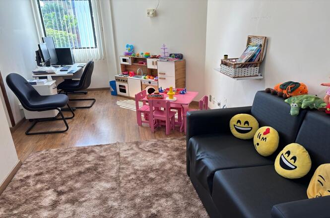 Foto horizontal: espaço lúdico com brinquedos infantis, mesa colorida e sofá com almofadas, ambiente acolhedor, tons claros.